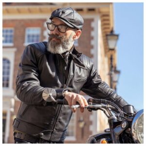 A bearded man wearing a leather jacket and a Belstaff Hislop Wax Cotton Cap on a motorcycle.