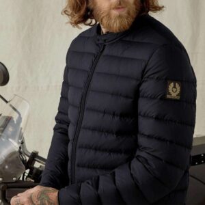 A man wearing the Belstaff Long Way Up-Down Jacket and a beard is standing next to a motorcycle.